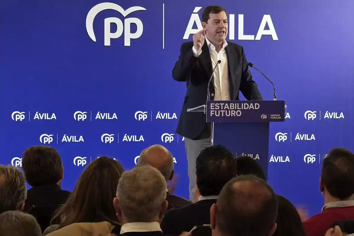 Un hombre de traje habla en un atril frente a un público en un evento del Partido Popular en Ávila con un fondo azul que muestra los logotipos del partido y las palabras estabilidad y futuro Un hombre de traje habla en un atril frente a un público en un evento del Partido Popular en Ávila con un fondo azul que muestra los logotipos del partido y las palabras estabilidad y futuro