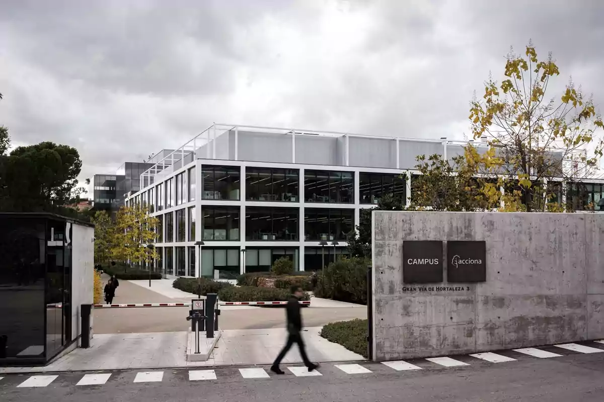 Edificio moderno de oficinas con fachada de vidrio y estructura blanca, rodeado de árboles y vegetación, con una persona cruzando la calle frente a la entrada principal donde hay un letrero que indica Campus Acciona