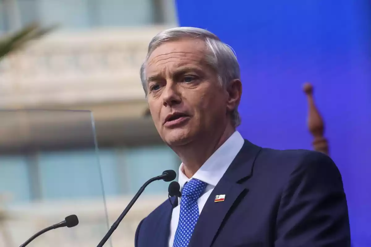 Hombre de cabello canoso con traje azul hablando frente a un micrófono en un escenario con fondo azul