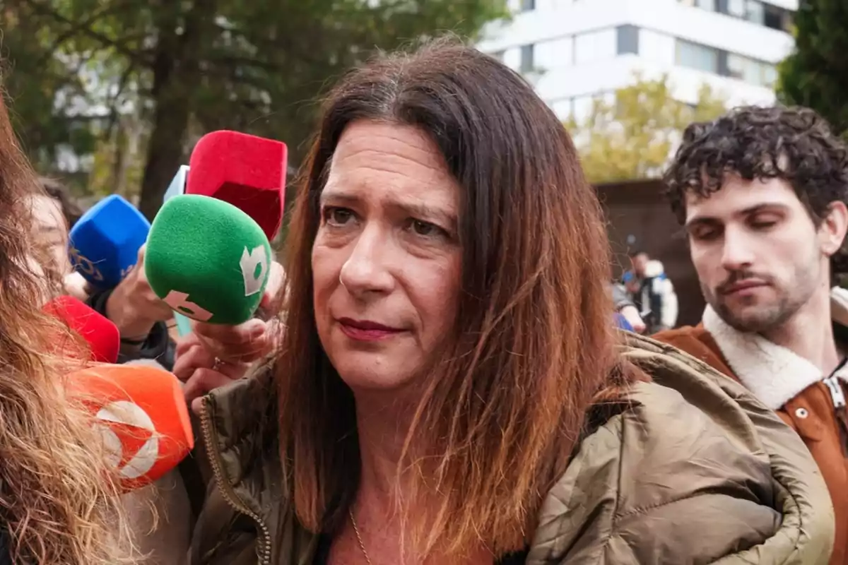Mujer de cabello castaño oscuro con expresión seria rodeada de micrófonos de colores mientras es entrevistada al aire libre