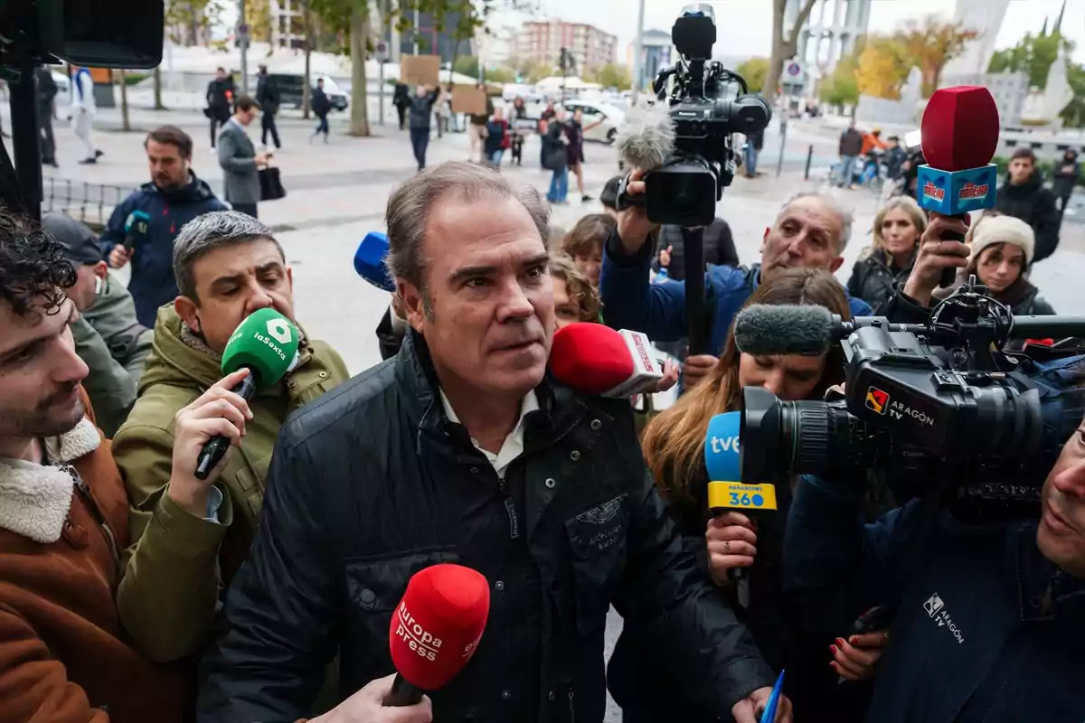 Un hombre con chaqueta oscura es rodeado por varios periodistas y cámaras de televisión mientras responde preguntas en la calle Un hombre con chaqueta oscura es rodeado por varios periodistas y cámaras de televisión mientras responde preguntas en la calle