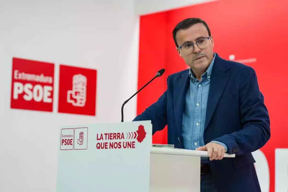 Hombre con gafas y chaqueta azul hablando en un atril con el logo de Extremadura PSOE y el lema La tierra que nos une sobre fondo rojo