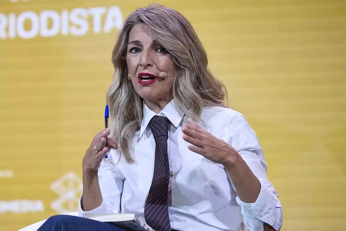 Mujer de cabello rubio y largo hablando en un evento con micrófono de diadema, camisa blanca y corbata oscura, sosteniendo un bolígrafo azul y un cuaderno frente a un fondo amarillo