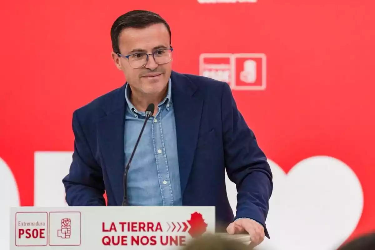Hombre con gafas y chaqueta azul hablando en un atril con fondo rojo y logotipos del PSOE
