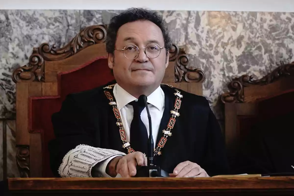 Hombre con gafas y toga sentado en una silla de madera tallada frente a un micrófono en un entorno formal