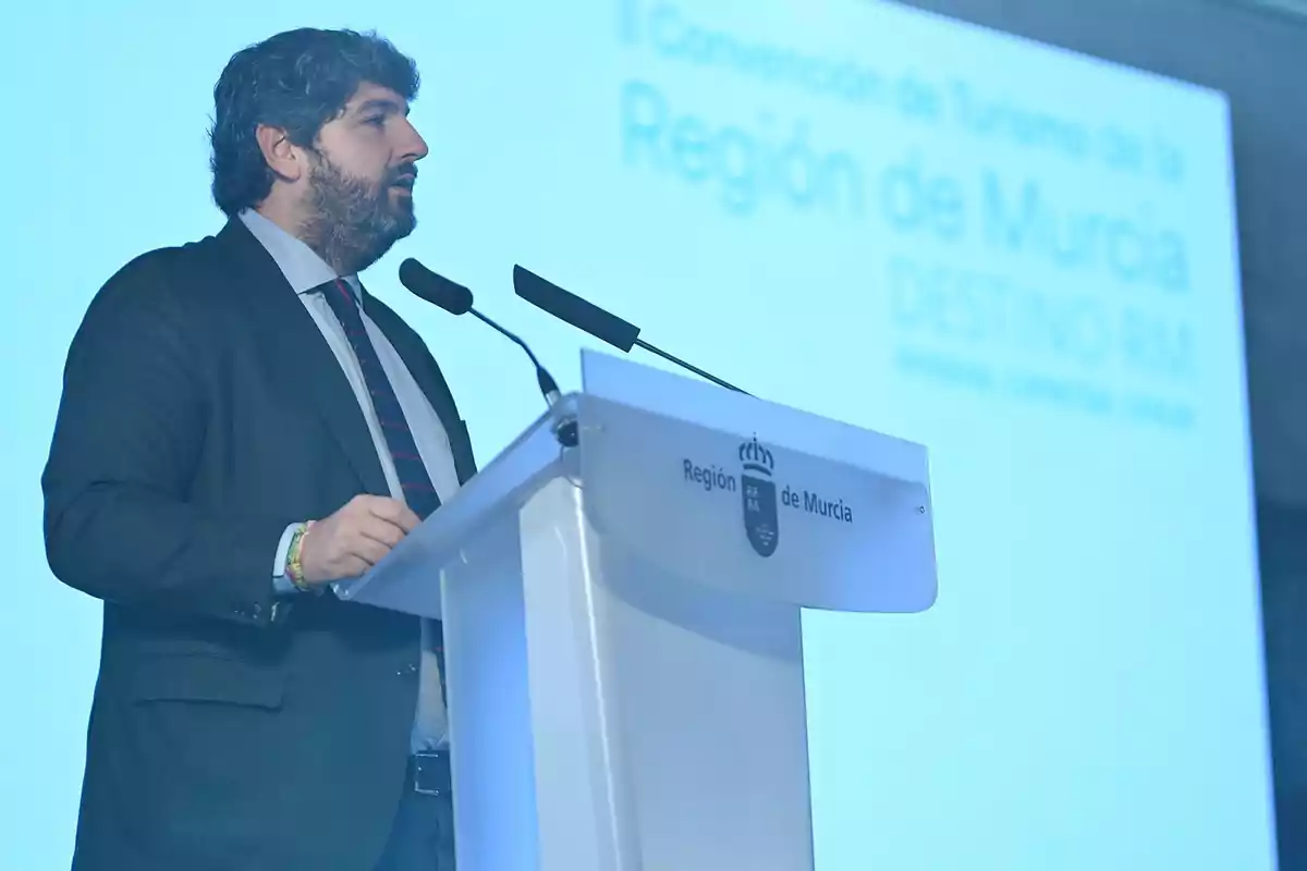 Hombre de traje hablando en un atril durante un evento de la Región de Murcia con un fondo azul y letras desenfocadas