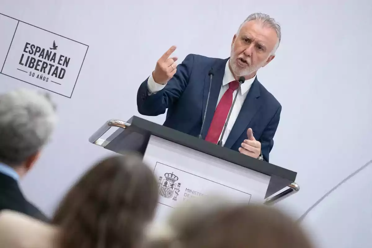 Hombre de cabello canoso y barba hablando en un atril con micrófonos durante un evento titulado España en libertad 50 años frente a una audiencia