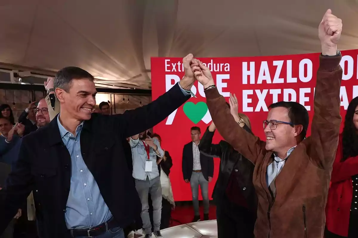 Dos hombres sonríen y levantan las manos entrelazadas en señal de celebración frente a un cartel rojo con mensajes políticos rodeados de personas aplaudiendo.