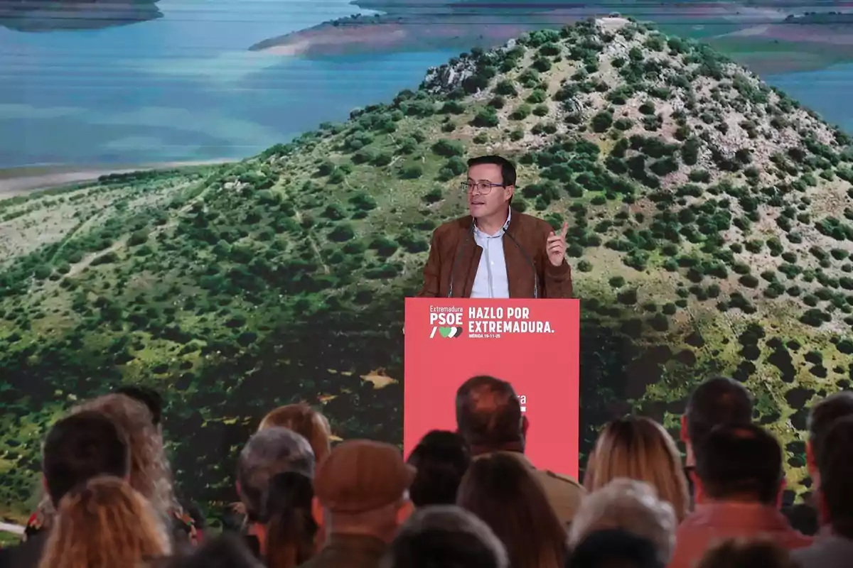 Un hombre da un discurso en un escenario frente a un público, con un fondo de paisaje natural y un atril rojo con el texto Hazlo por Extremadura y el logo del PSOE. Un hombre da un discurso en un escenario frente a un público, con un fondo de paisaje natural y un atril rojo con el texto Hazlo por Extremadura y el logo del PSOE.