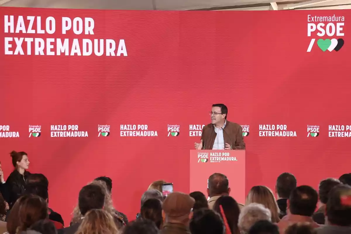 Un hombre da un discurso en un mitin del PSOE de Extremadura frente a un público, con un fondo rojo que tiene el lema Hazlo por Extremadura. Un hombre da un discurso en un mitin del PSOE de Extremadura frente a un público, con un fondo rojo que tiene el lema Hazlo por Extremadura.