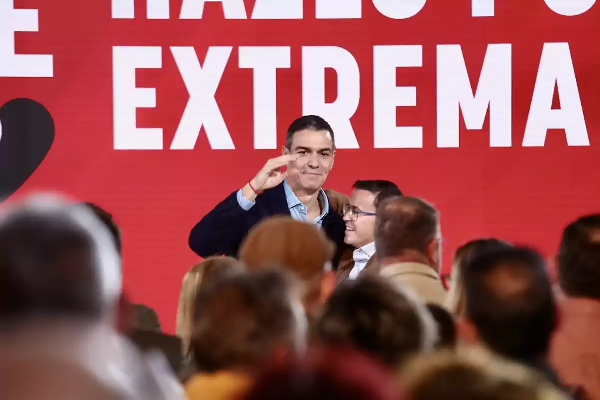 Un hombre saluda a una multitud en un evento con fondo rojo y letras grandes blancas Un hombre saluda a una multitud en un evento con fondo rojo y letras grandes blancas