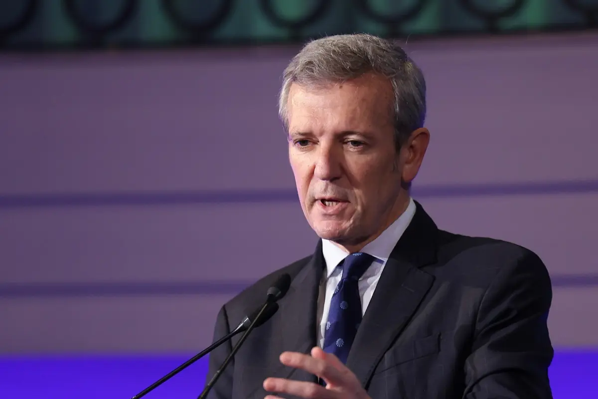 Hombre de cabello canoso con traje oscuro hablando frente a un micrófono en un escenario iluminado con luces moradas