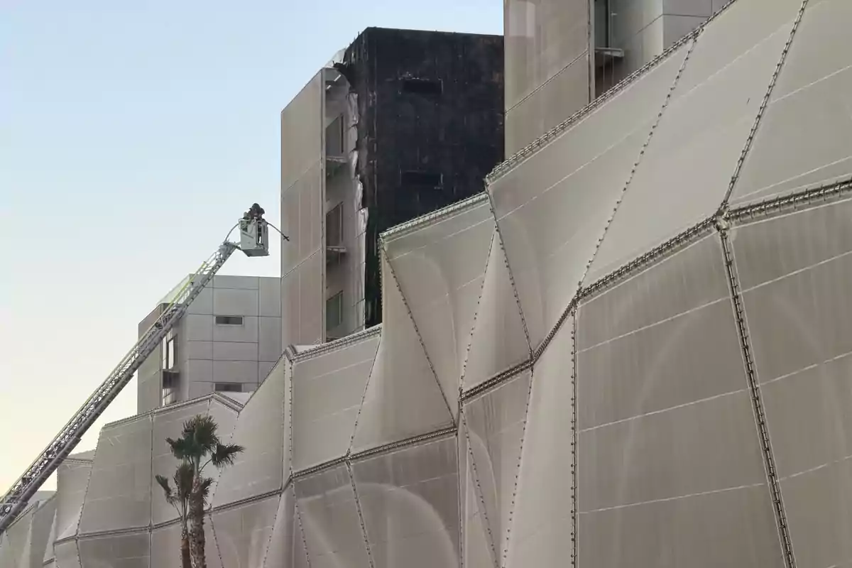 Un bombero en una grúa apaga un incendio en un edificio moderno con una fachada geométrica y parte de la estructura quemada