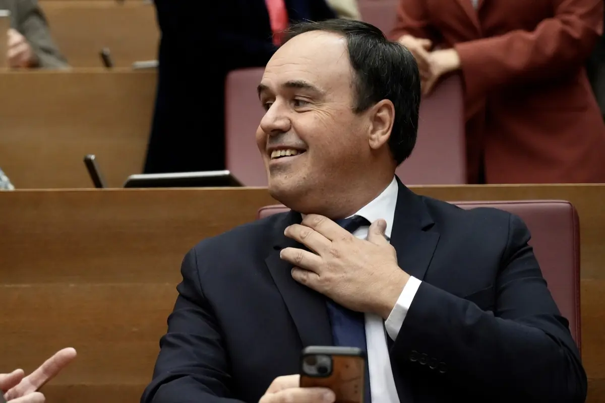 Hombre de traje oscuro sonriendo mientras sostiene un teléfono móvil y se ajusta la corbata en una sala de reuniones