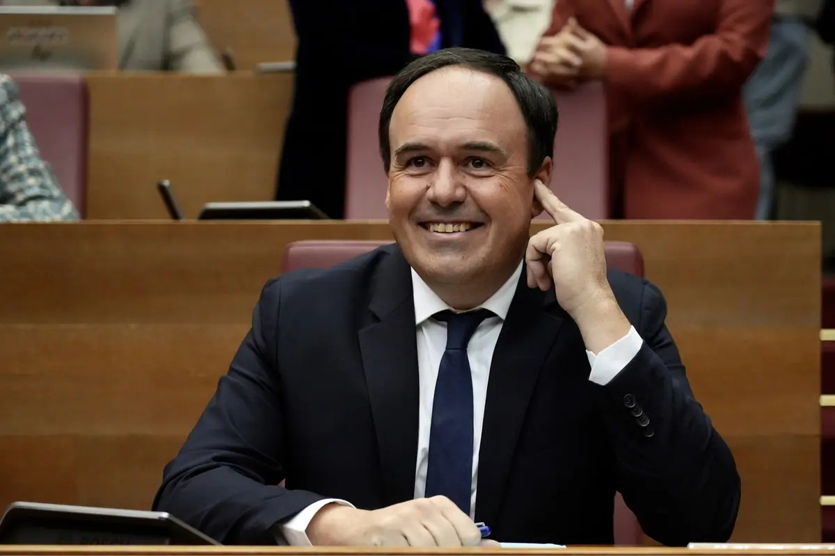 Hombre de traje oscuro y corbata azul sonriendo mientras está sentado en una sala de reuniones