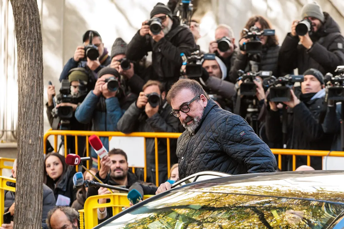 Un hombre con gafas y chaqueta negra sale de un coche mientras numerosos periodistas y fotógrafos lo rodean y le toman fotos frente a una valla amarilla