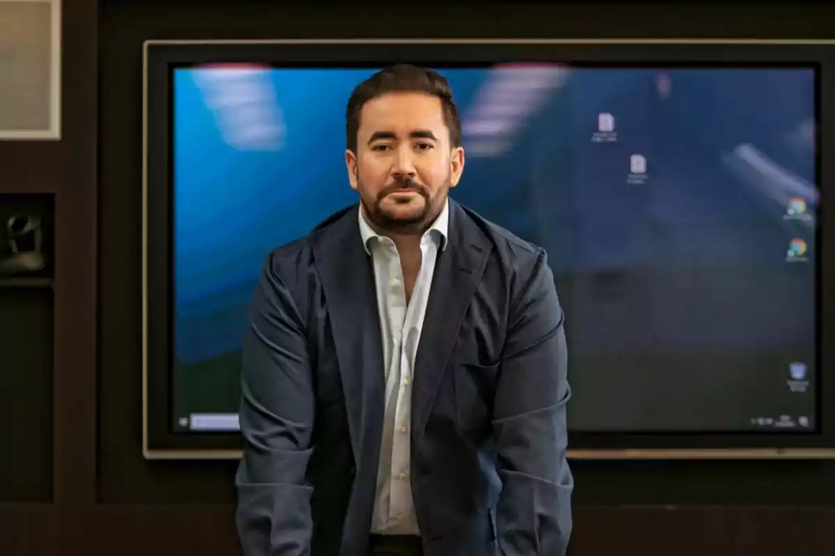 Hombre de barba y cabello corto con saco azul marino y camisa blanca de pie frente a una pantalla grande en una oficina moderna