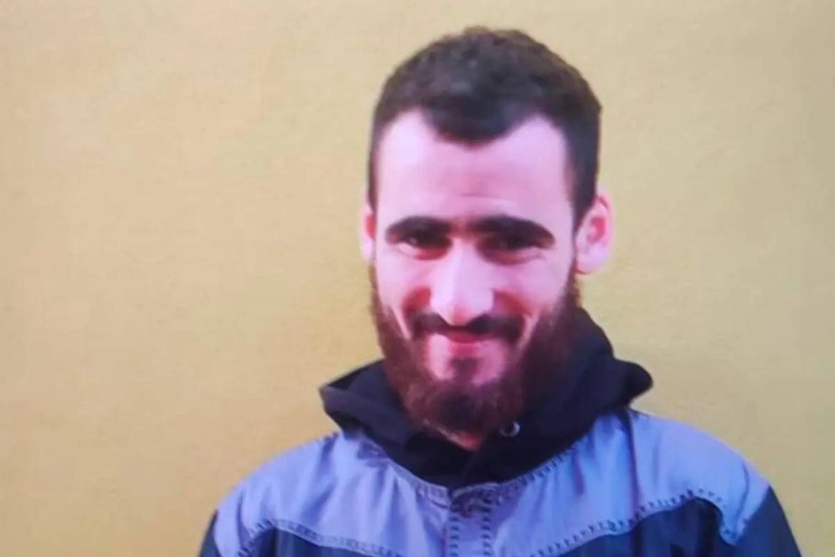 Hombre joven con barba y cabello corto, sonriendo frente a una pared amarilla, usando una chaqueta gris y negra.