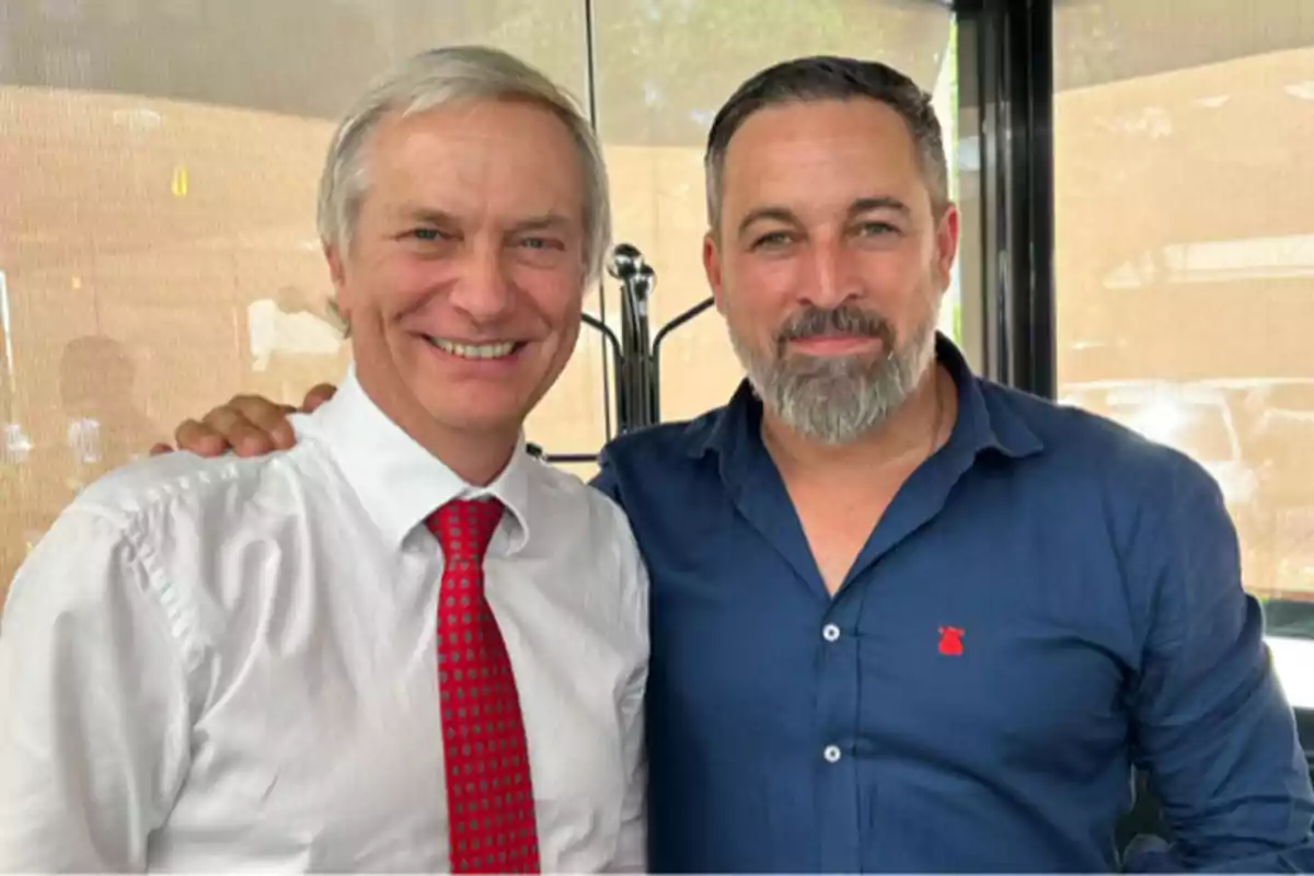 Dos hombres posan juntos y sonríen frente a la cámara, uno lleva camisa blanca y corbata roja y el otro viste camisa azul