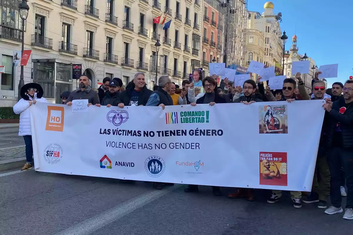 Un grupo de personas marcha por una calle sosteniendo una pancarta grande que dice Las víctimas no tienen género y Violence has no gender, rodeados de edificios y banderas, mientras otros manifestantes al fondo levantan carteles con corazones dibujados.