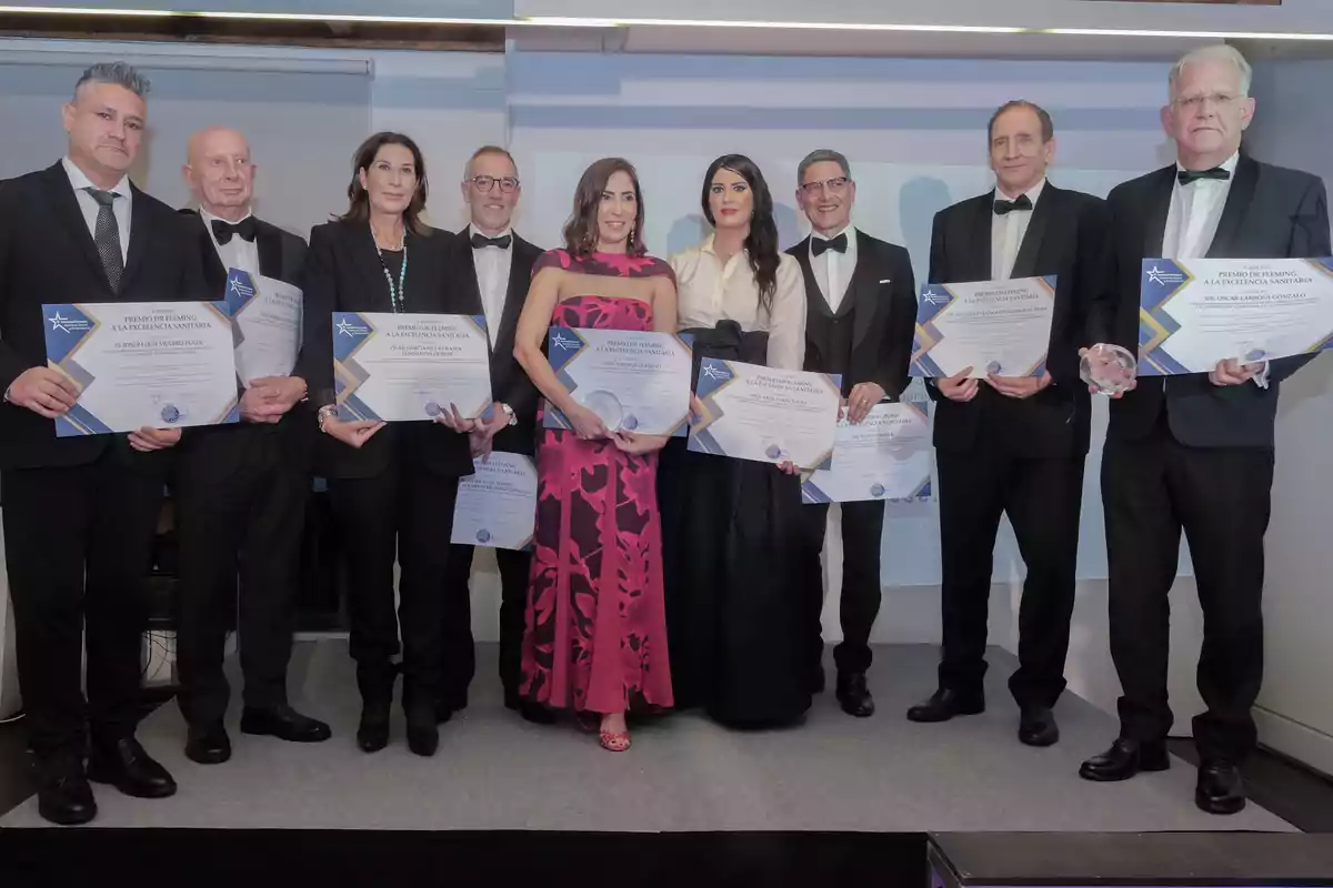 Grupo de personas elegantemente vestidas posando en un escenario mientras sostienen diplomas de reconocimiento