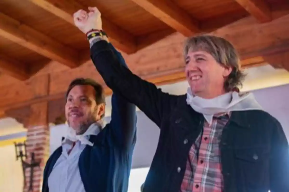 Dos hombres sonríen y levantan los brazos entrelazados en señal de celebración dentro de un ambiente con techo de madera