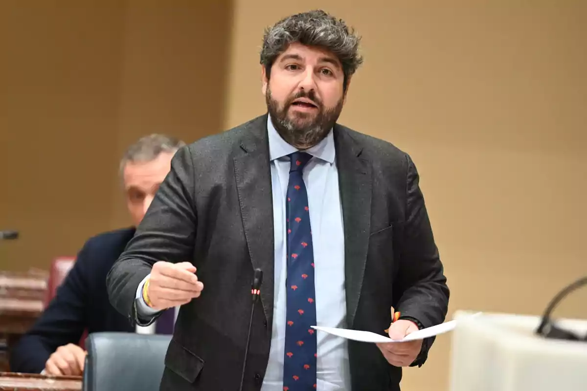 Hombre de cabello rizado y barba hablando en un recinto formal mientras sostiene unos papeles