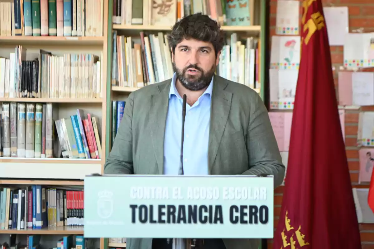 Hombre de cabello rizado y barba hablando en un atril con el mensaje contra el acoso escolar tolerancia cero en una biblioteca con estanterías de libros y una bandera roja al fondo