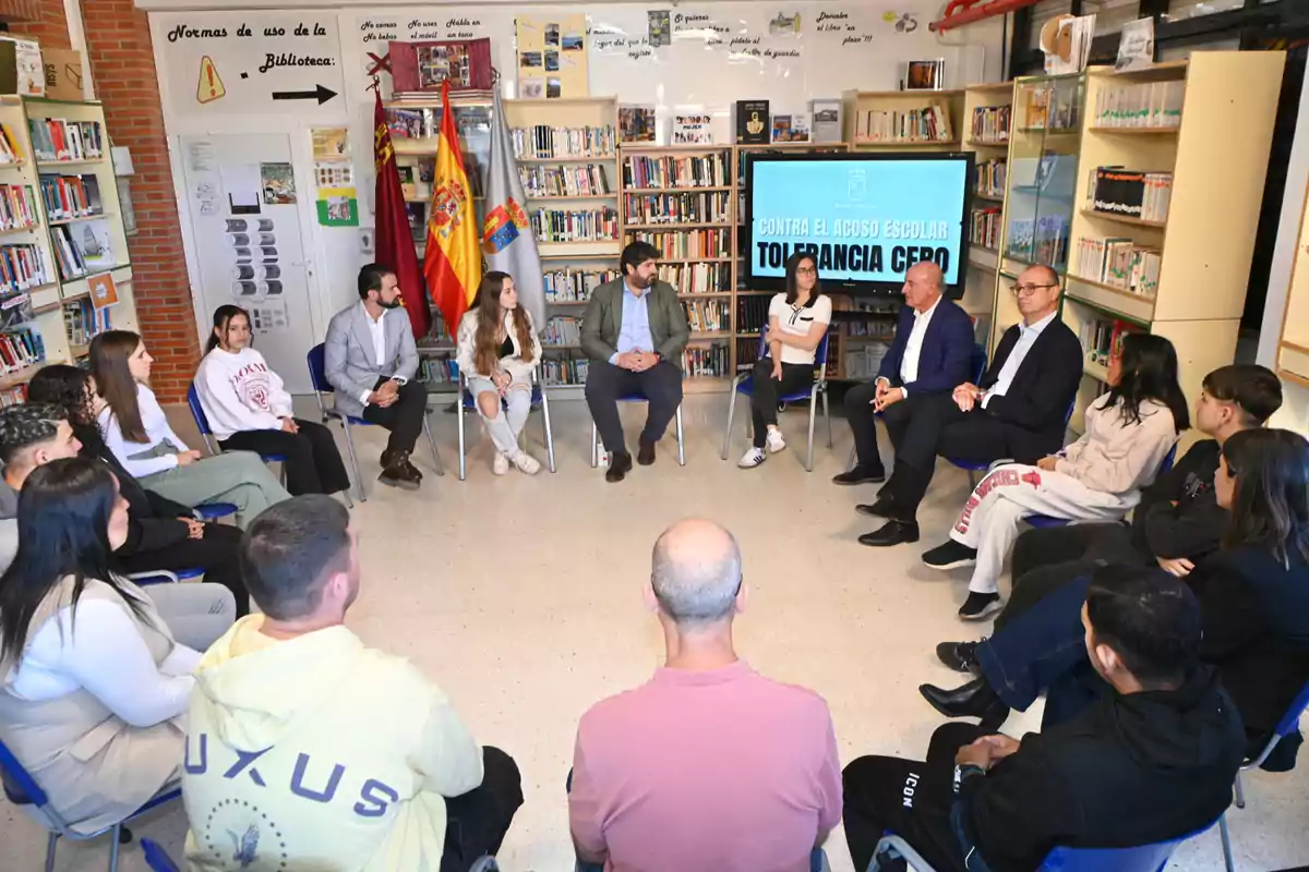 Un grupo de personas sentadas en círculo en una biblioteca escolar participando en una charla sobre la tolerancia cero al acoso escolar, con una pantalla de fondo que muestra el mensaje contra el acoso y varias banderas junto a estanterías llenas de libros.