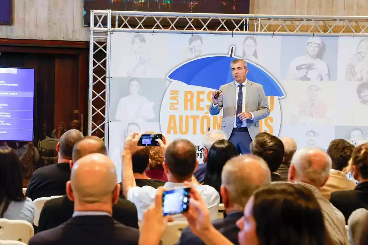 Un hombre de traje da una presentación frente a una audiencia mientras varias personas toman fotos con sus teléfonos y detrás de él hay un cartel con un paraguas azul y letras grandes.