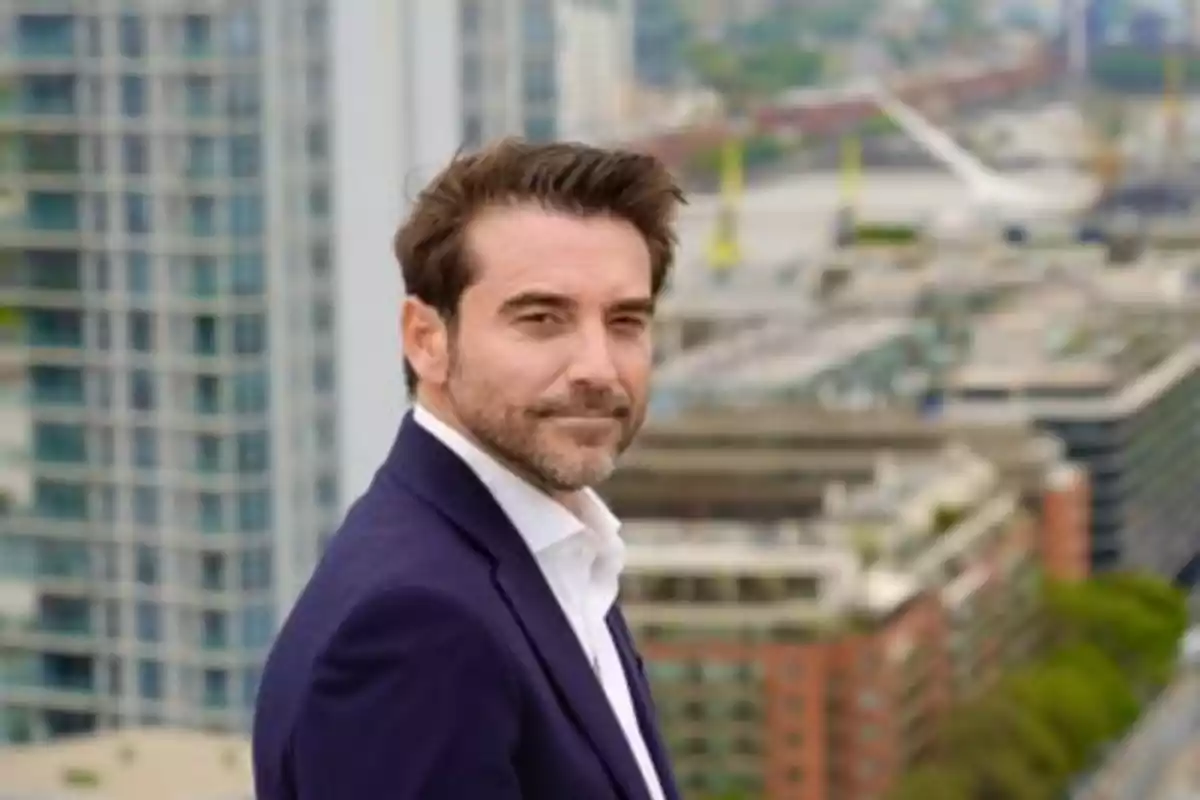 Hombre de cabello castaño y barba con saco azul posando en una terraza con edificios de fondo