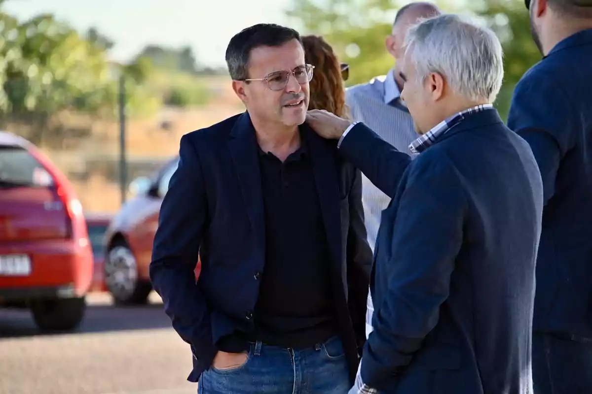 Dos hombres conversan al aire libre mientras uno de ellos apoya la mano en el hombro del otro, ambos visten ropa formal y hay autos estacionados al fondo.