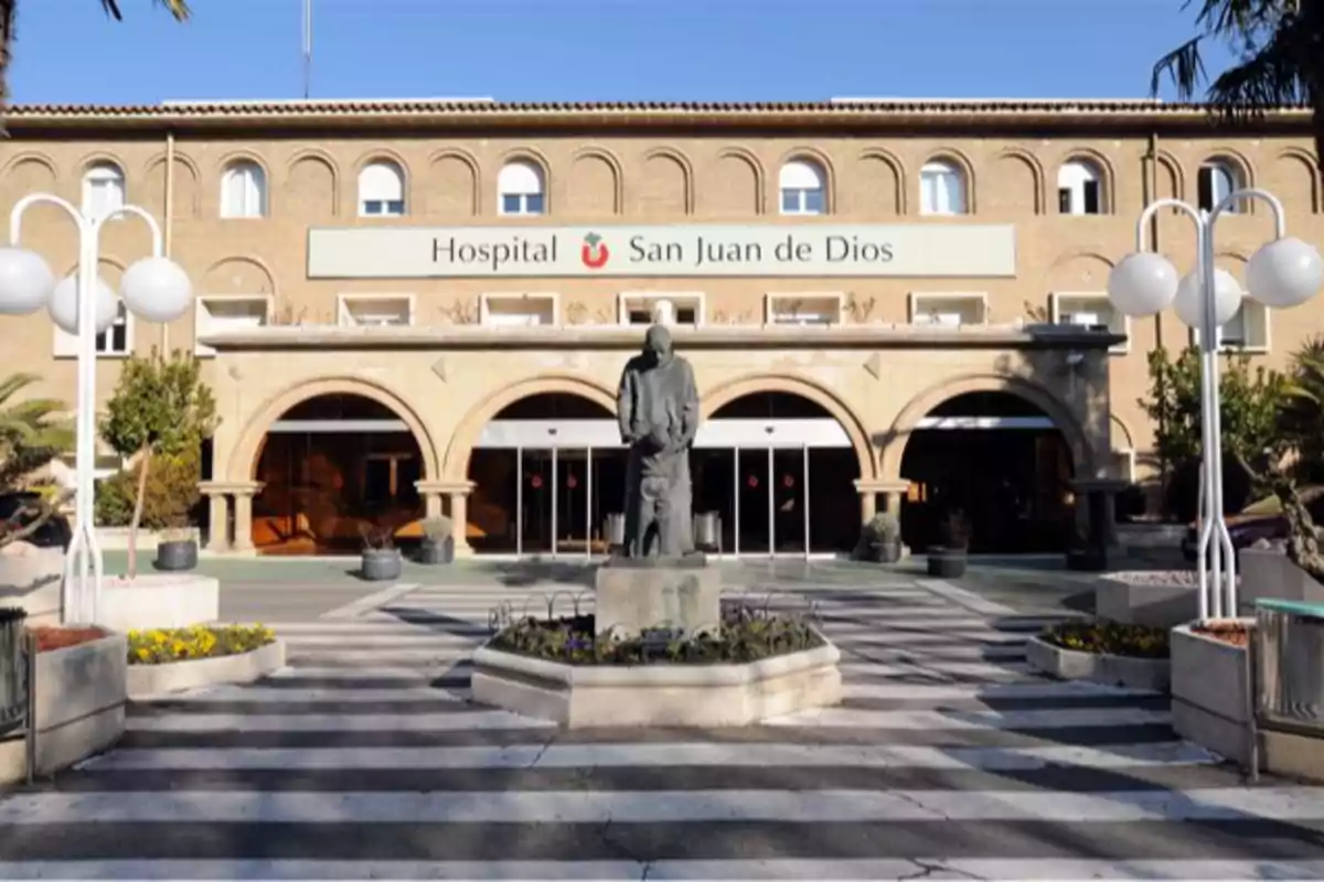 Fachada principal de un hospital con una estatua en el centro y un paso de peatones en primer plano