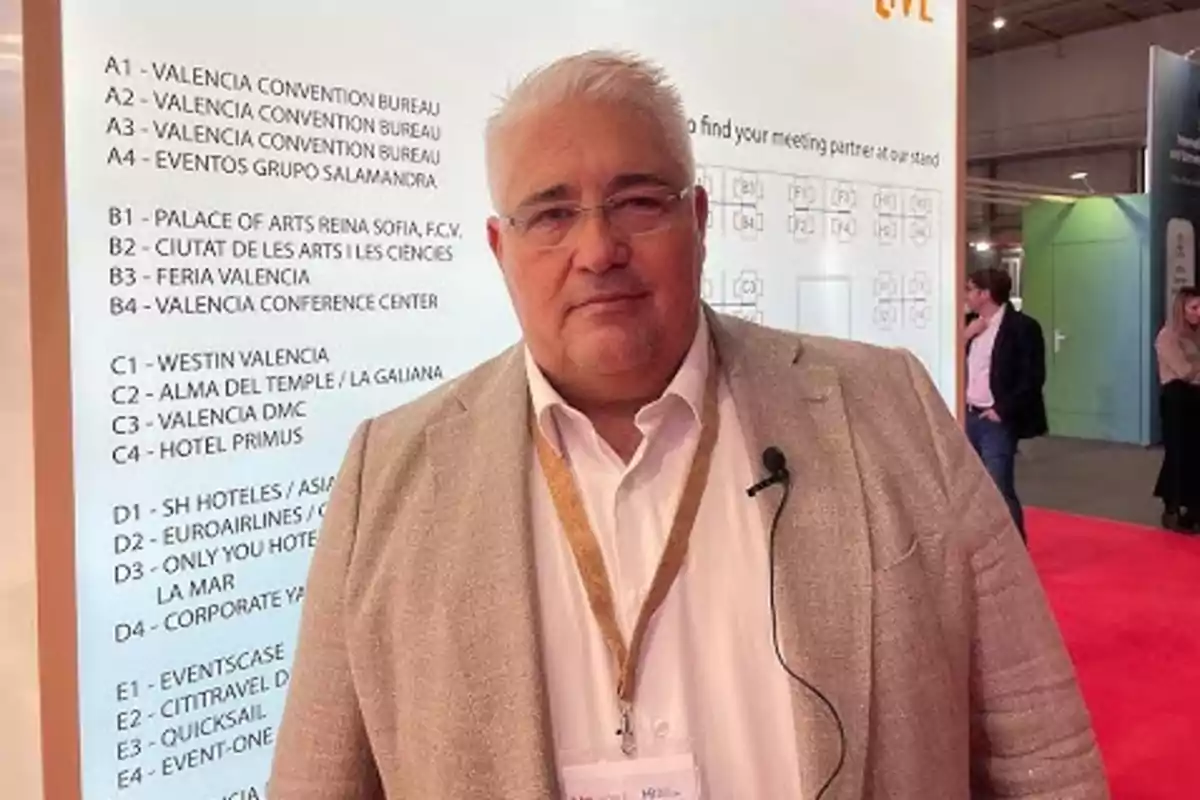 Hombre de cabello canoso y gafas con chaqueta beige y camisa blanca posando en un evento profesional con fondo de cartel informativo y alfombra roja