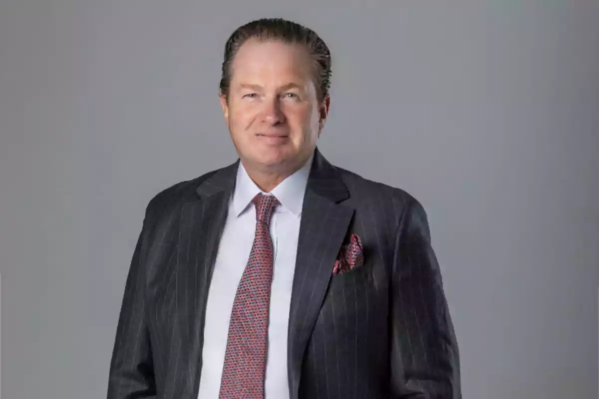 Hombre de mediana edad con cabello corto y peinado hacia atrás, usando traje oscuro con camisa blanca y corbata estampada, posando frente a un fondo gris.