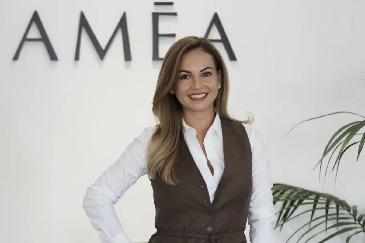 Mujer sonriente de cabello largo y rubio vestida con camisa blanca y chaleco marrón posando frente a una pared blanca con letras grandes y una planta a un lado