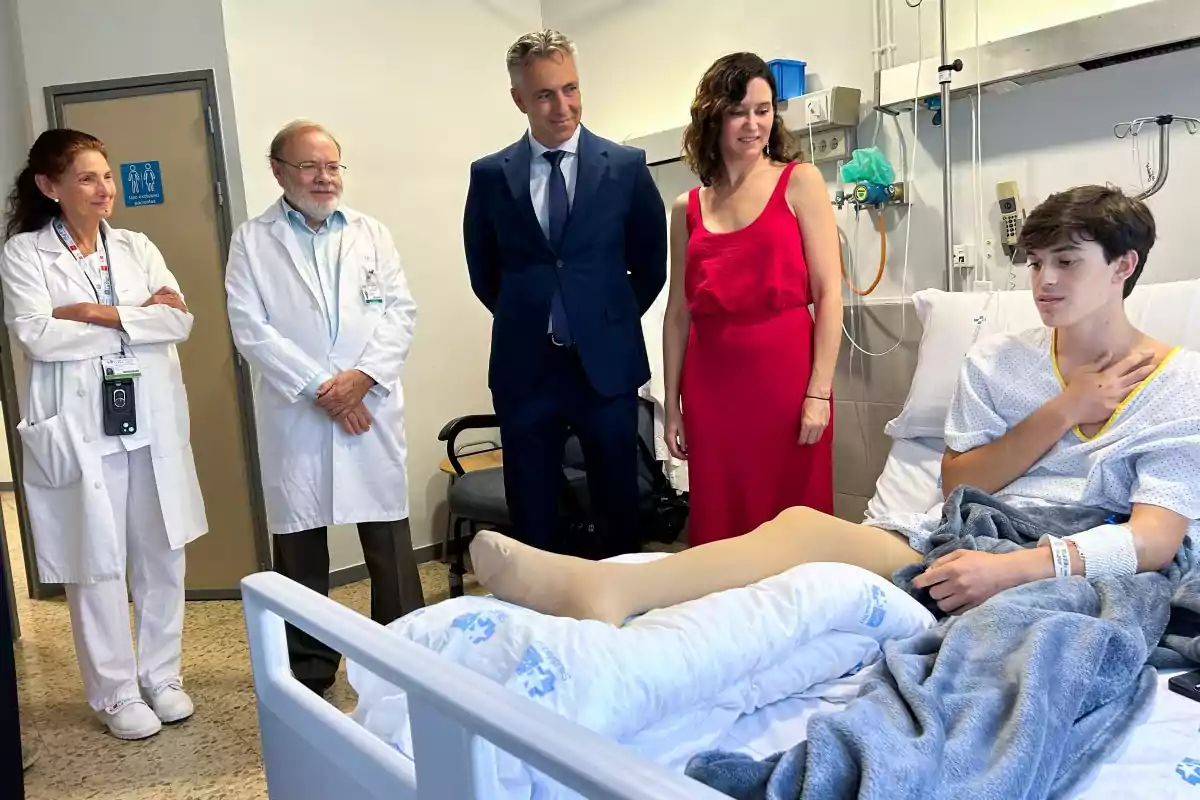 Un joven paciente está acostado en una cama de hospital con una pierna vendada mientras dos médicos, un hombre de traje y una mujer con vestido rojo lo visitan y conversan con él Un joven paciente está acostado en una cama de hospital con una pierna vendada mientras dos médicos, un hombre de traje y una mujer con vestido rojo lo visitan y conversan con él