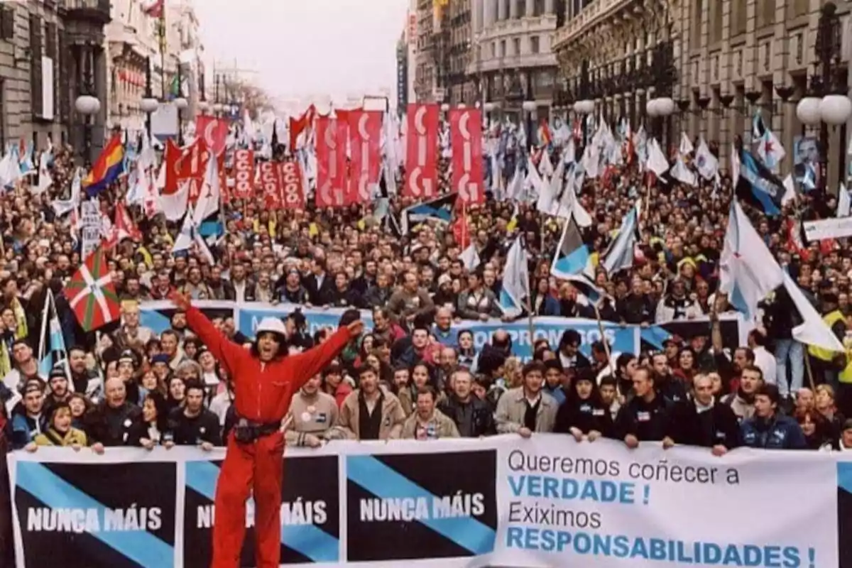 Multitud de personas marchando por una avenida con pancartas y banderas, al frente se lee Nunca Máis y un hombre de rojo con los brazos abiertos destaca en primer plano