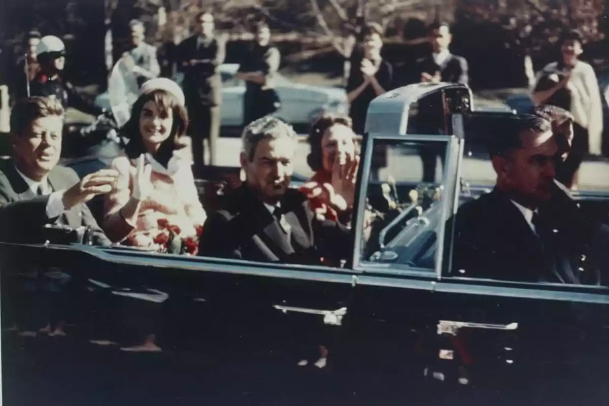 Desfile en automóvil descapotable con Kennedy y Jaqueline saludando al público mientras avanzan por una calle escoltados por motociclistas y observados por transeúntes
