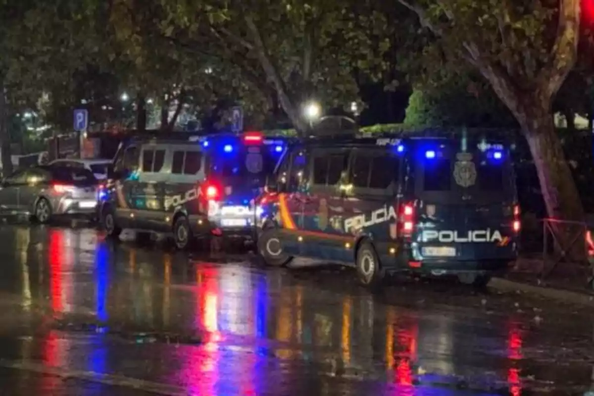 Varios vehículos de la policía con las luces encendidas están estacionados en una calle mojada durante la noche, reflejando sus luces en el asfalto