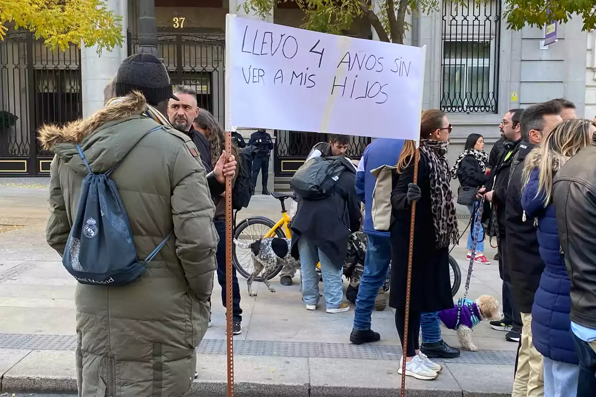 Un grupo de personas está reunido en la calle y una de ellas sostiene un cartel que dice que lleva cuatro años sin ver a sus hijos.