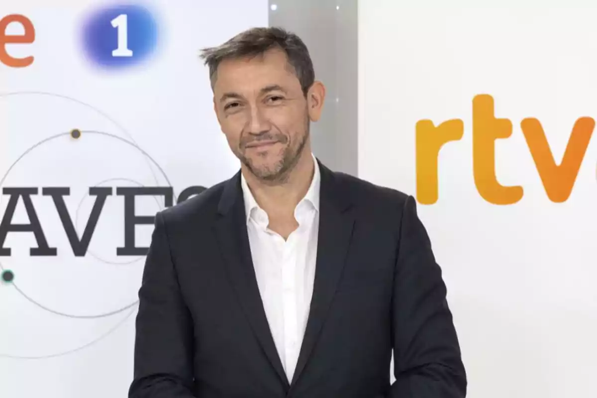 Hombre de cabello corto y barba con traje oscuro y camisa blanca posando frente a un fondo con logotipos de RTVE y un cartel de un programa televisivo Hombre de cabello corto y barba con traje oscuro y camisa blanca posando frente a un fondo con logotipos de RTVE y un cartel de un programa televisivo