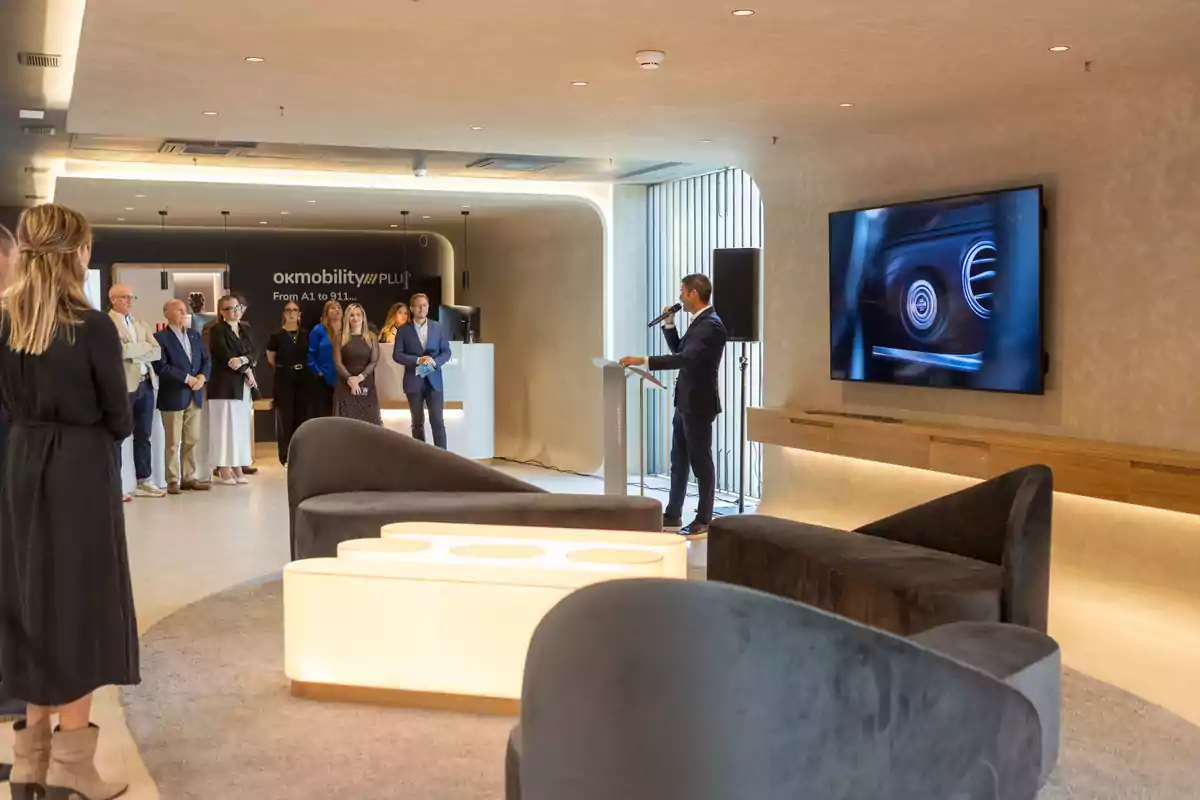 Un hombre de traje habla en un micrófono frente a un grupo de personas en una sala moderna con sofás y una pantalla grande en la pared