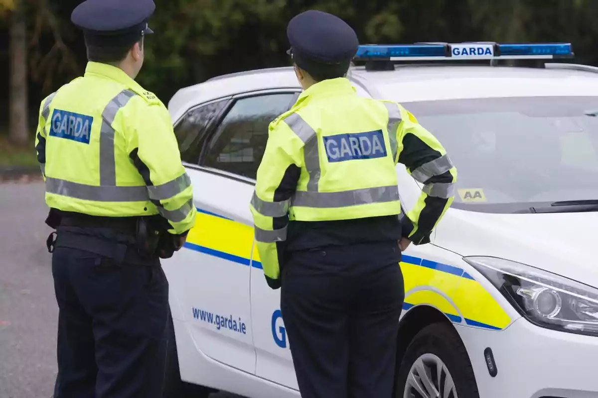 Dos agentes de policía irlandesa con chaquetas reflectantes de color amarillo junto a un coche patrulla blanco