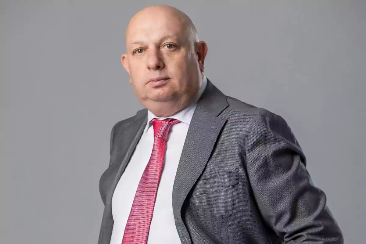 Hombre calvo con traje gris, camisa blanca y corbata roja posando frente a un fondo gris