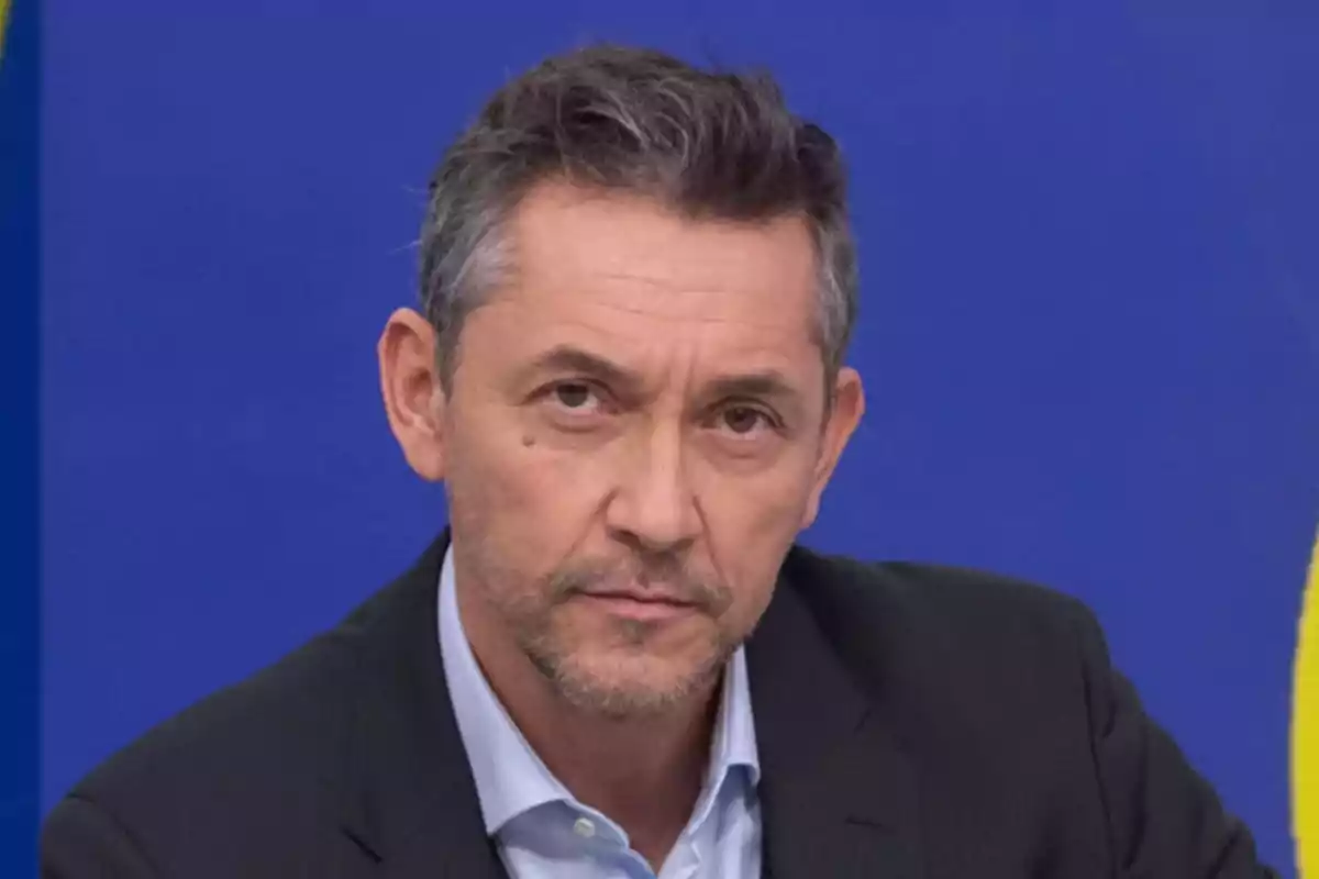 Hombre de cabello corto y canoso con barba ligera vistiendo saco oscuro y camisa clara frente a un fondo azul Hombre de cabello corto y canoso con barba ligera vistiendo saco oscuro y camisa clara frente a un fondo azul