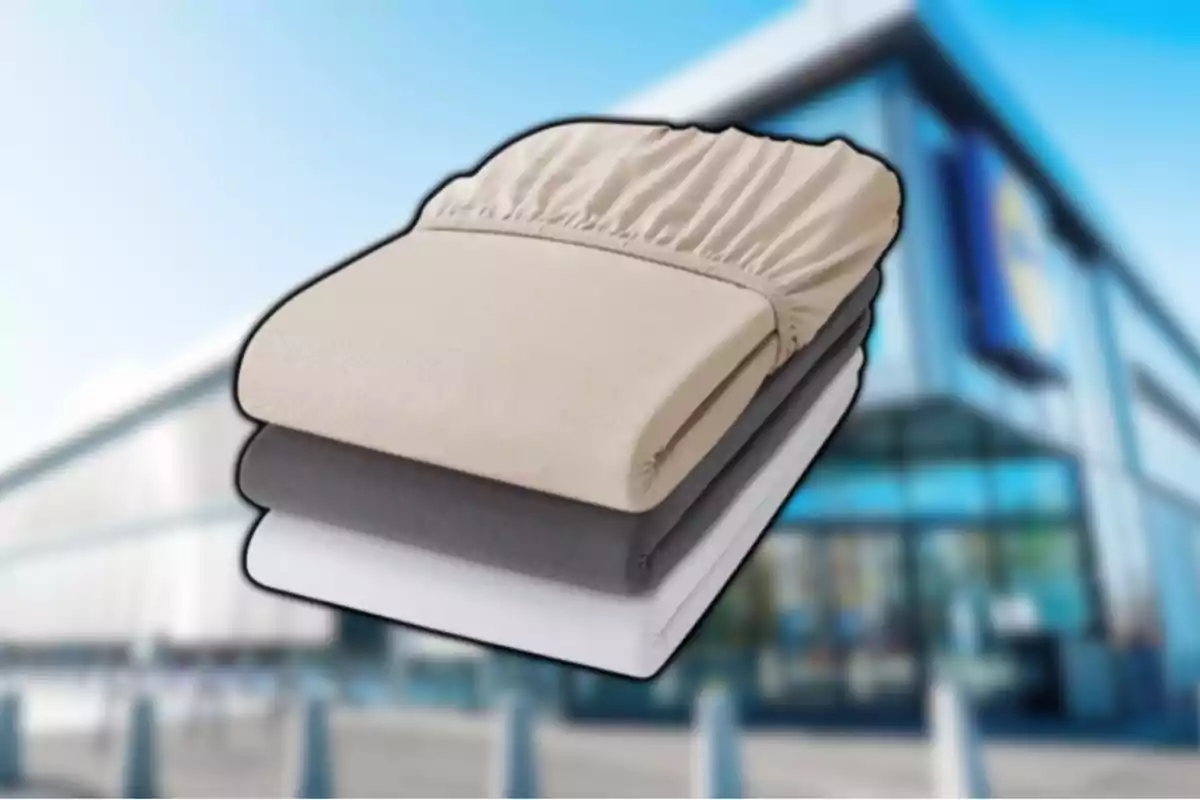 Tres sábanas ajustables en colores beige, gris y blanco apiladas frente a un edificio moderno desenfocado. Tres sábanas ajustables en colores beige, gris y blanco apiladas frente a un edificio moderno desenfocado.