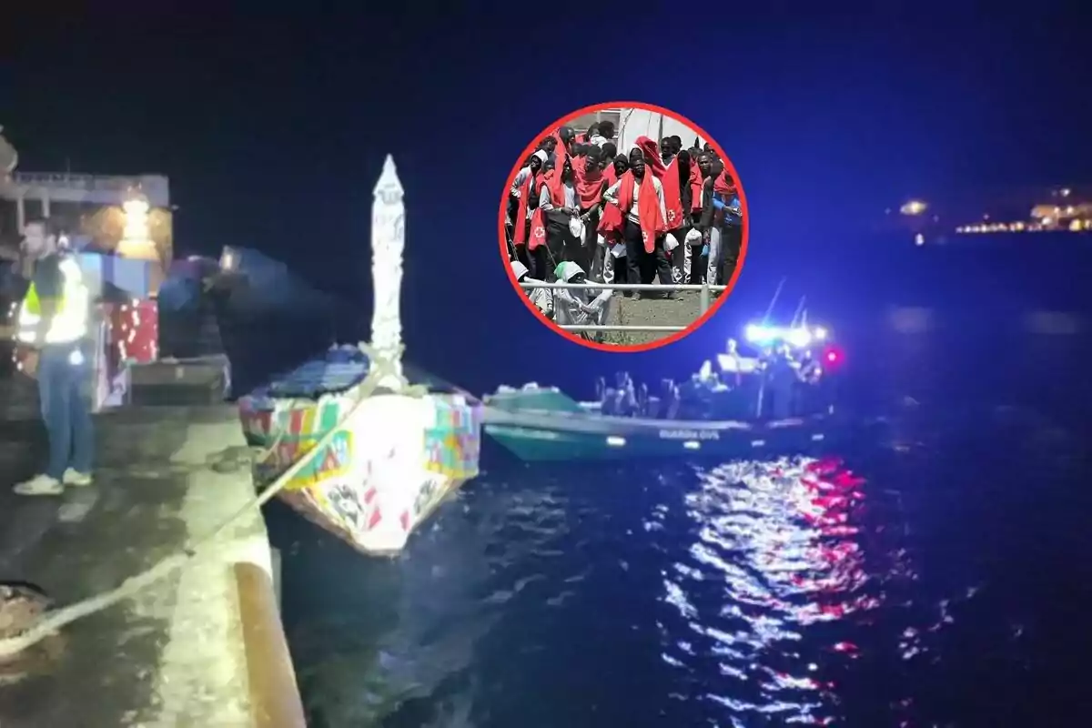 Un barco colorido amarrado en un muelle de noche con una lancha de la Guardia Civil en el agua y un grupo de personas con mantas rojas en un círculo superpuesto