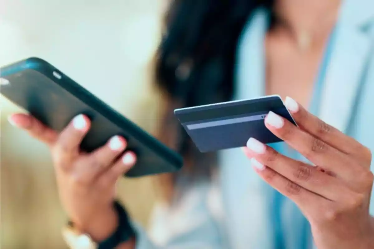 Persona sosteniendo una tarjeta bancaria en una mano y un teléfono móvil en la otra mientras realiza una compra en línea