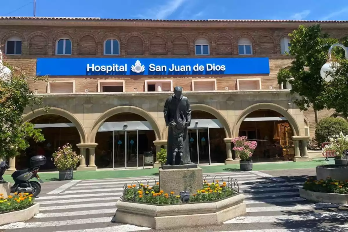 Fachada de un hospital con un letrero azul que dice Hospital San Juan de Dios y una estatua en el centro rodeada de flores y árboles Fachada de un hospital con un letrero azul que dice Hospital San Juan de Dios y una estatua en el centro rodeada de flores y árboles